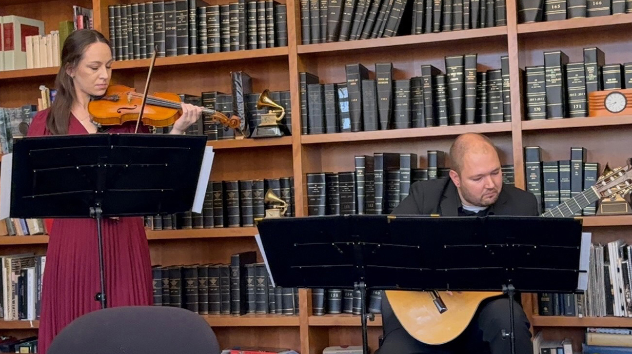 A young woman plays violin and a young man plays guitar in front of bookcases full of books.