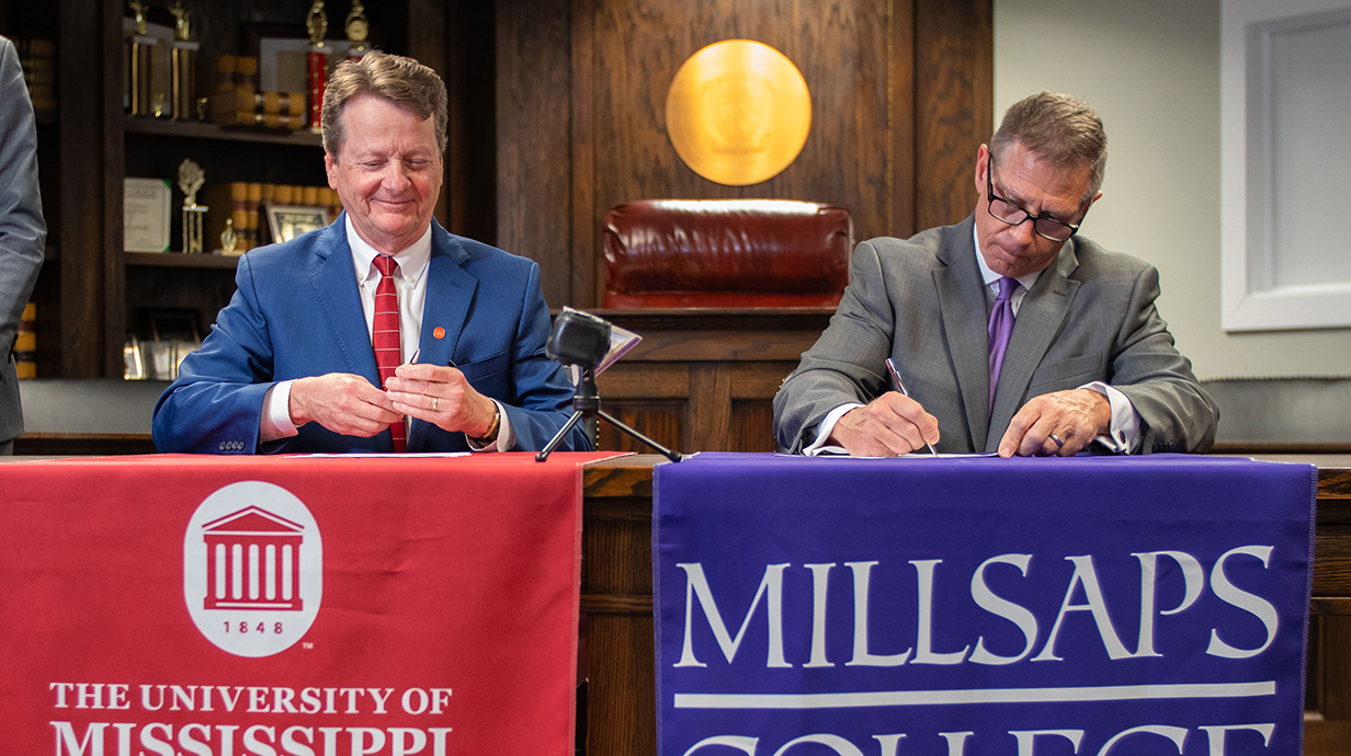 Two men sign papers while seated side-by-side at a table draped with banners.