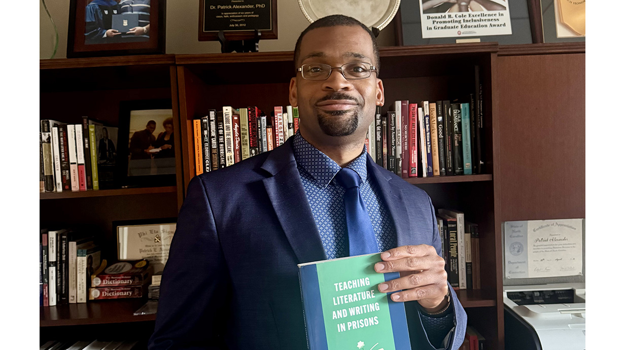 A man wearing a suit holds a book in front of full-length bookcases full of volumes.