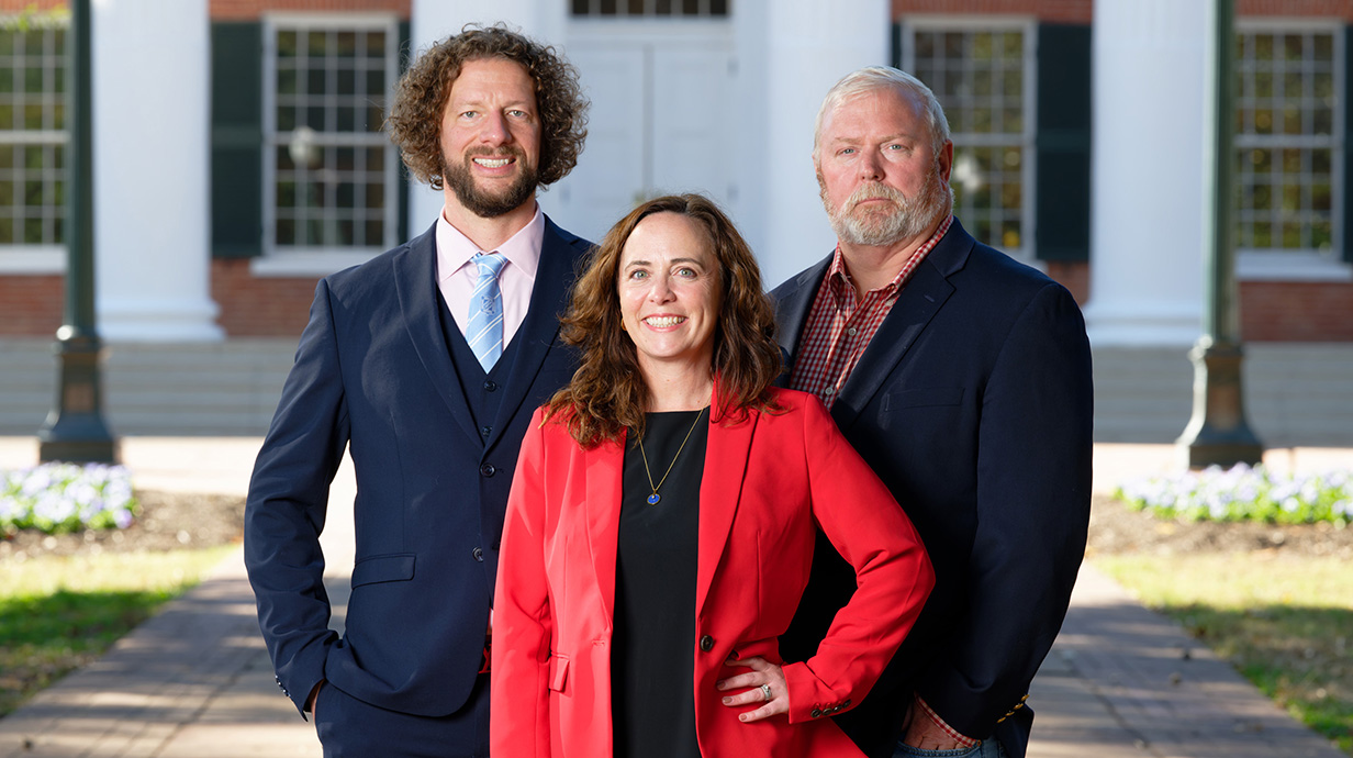 Two men and a woman stand outside a large red brick building with white columns.
