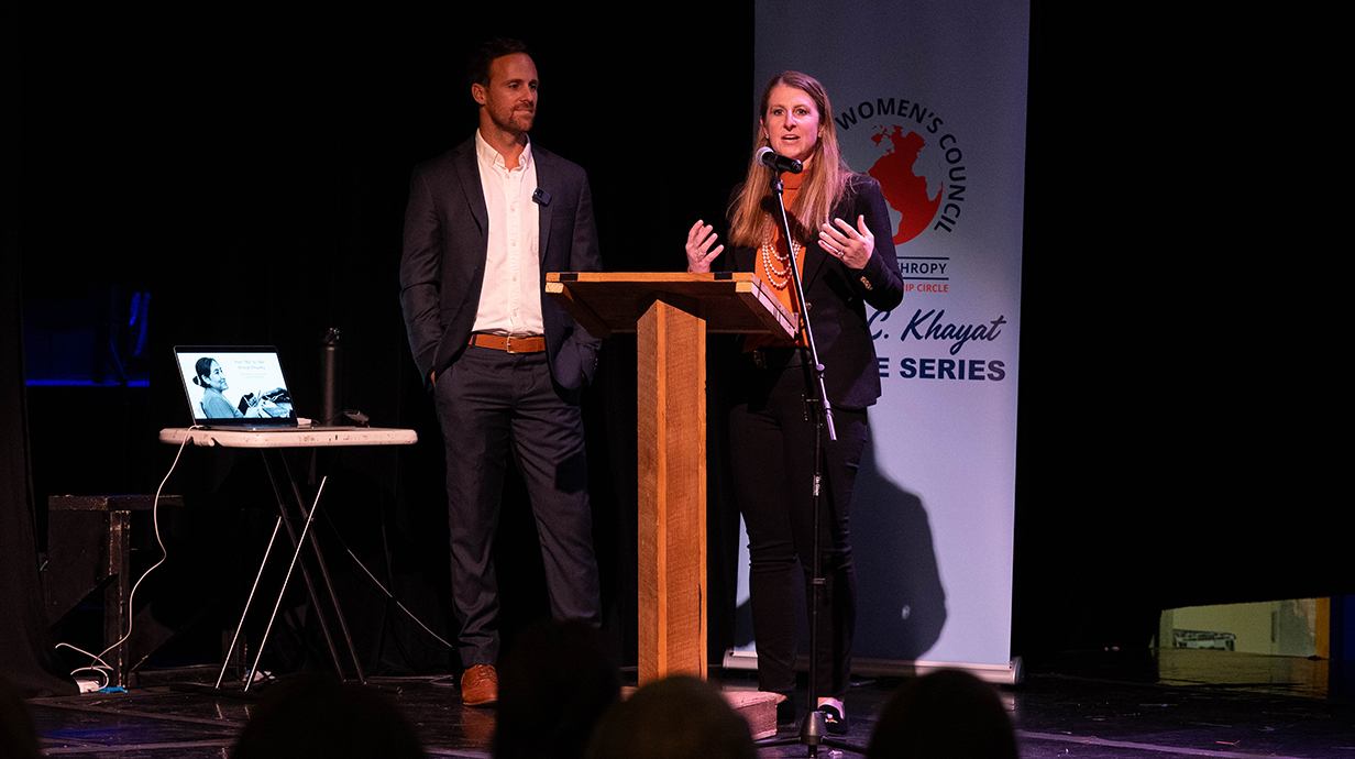 A woman speaks at a podium next to a young man.
