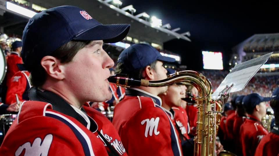 A young woman plays saxophone as part of a marching band in a stadium.