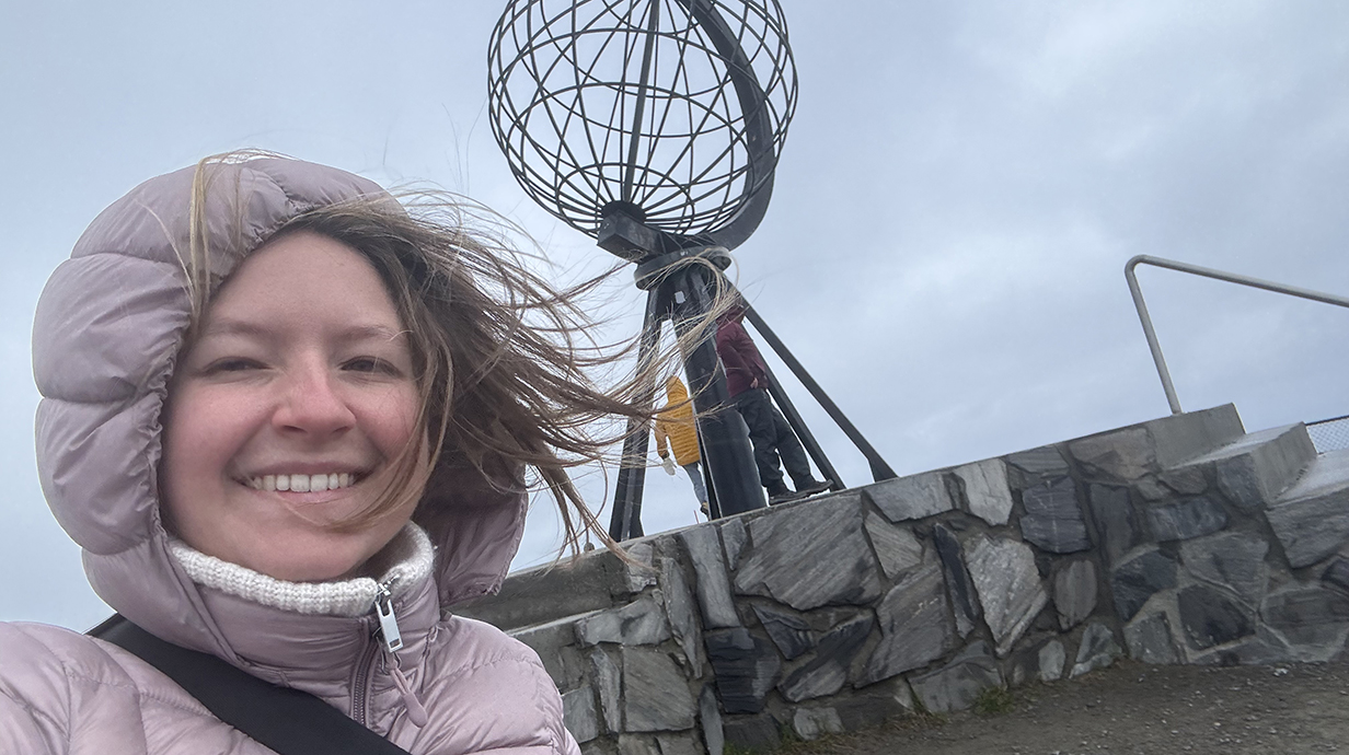A young woman wearing a pink coat and hood stands in front of a tower on a windy day.