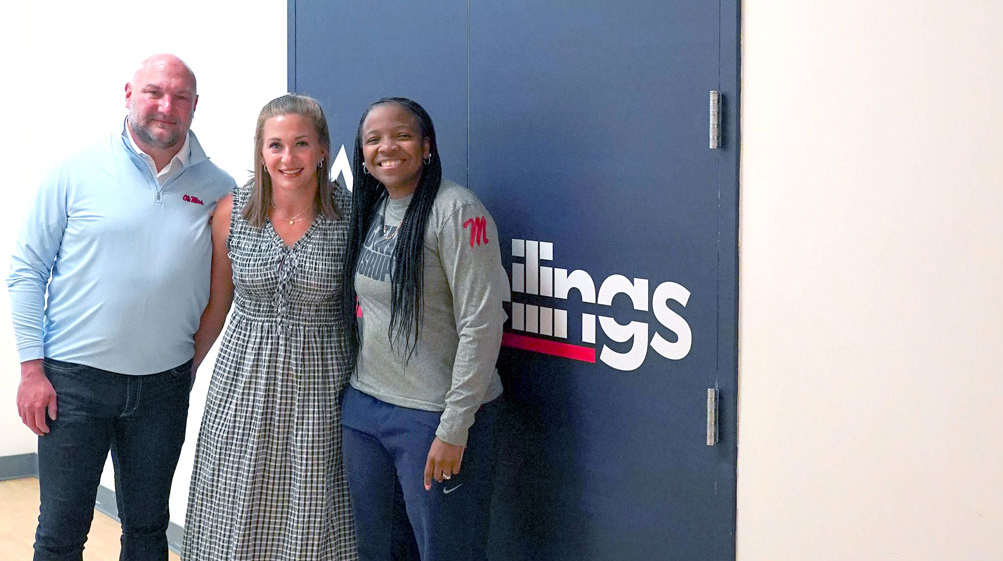 Two women and a man stand in a hallway just outside large blue double doors.