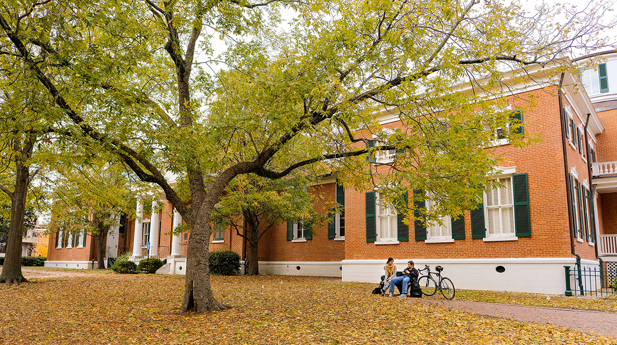 A couple sits in front of a red brick antebellum building with fall leaves scattered across the front lawn.