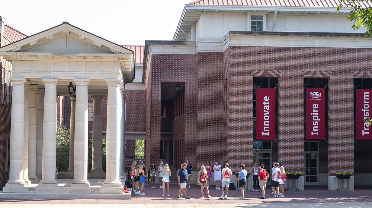 A group of young people walks in front of a large red brick building with red banners on its facade.
