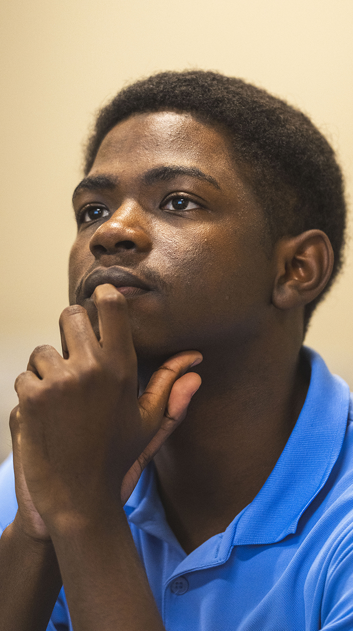 A young man wearing a blue polo shirt holds his chin while he looks at a speaker.