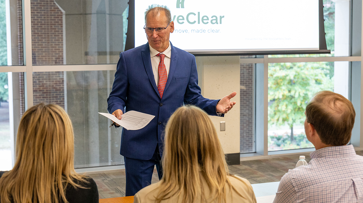 A man standing in the front of a conference hall speaks to two seated women and a seated man.