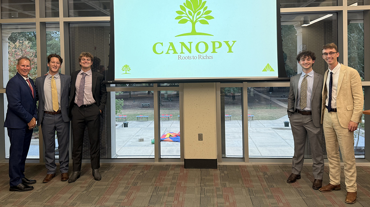 Four young men and a man, all wearing suits, stand around a projection screen reading 'Canopy.'