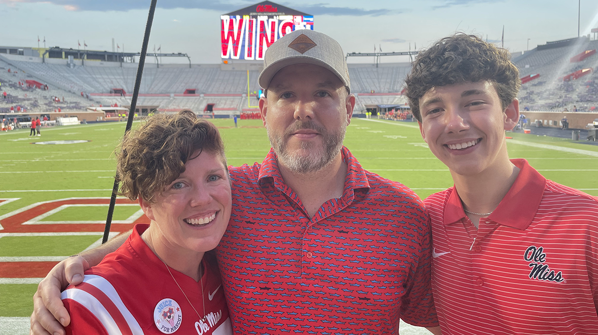 A woman and two men, all wearing Ole Miss apparel, stand in a nearly empty stadium after a football game.