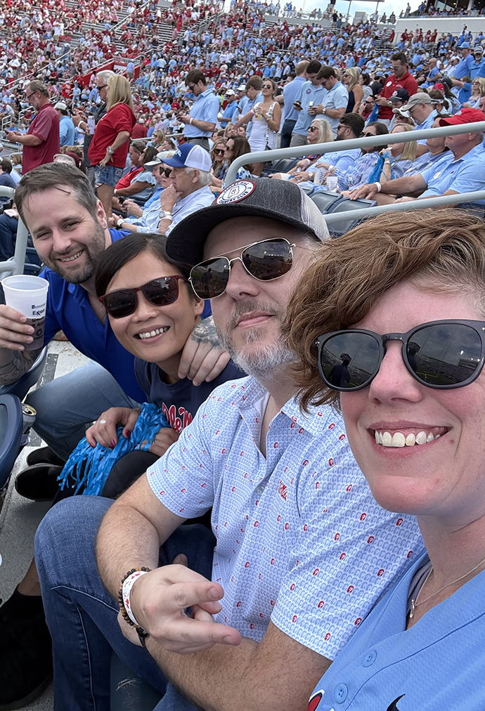 Two men and two women take a selfie while seated in a crowded football stadium.
