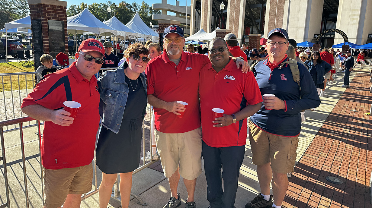 A group of people wearing Ole Miss gear stand on a concrete plaza outside a football stadium.