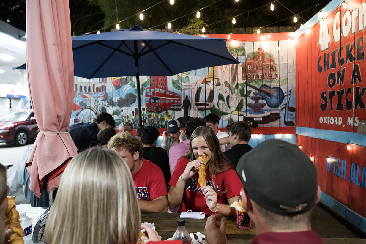 A group of people sit at picnic tables under string lights outside 4 Corners Chevron. A young woman in a red shirt eats a chicken on a stick while talking with others.