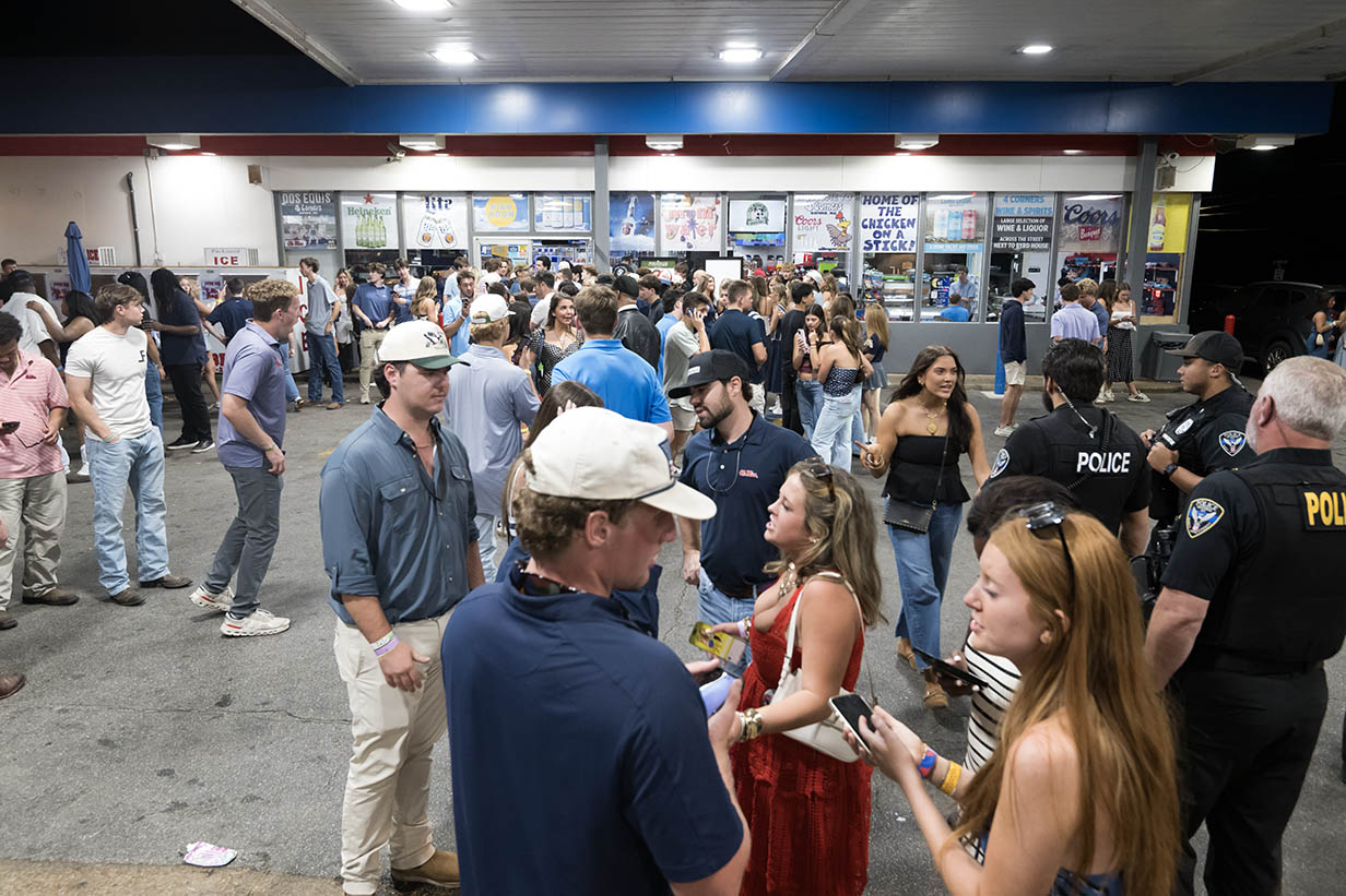 A large crowd lines up outside 4 Corners Chevron at night, with police officers standing nearby as people wait to enter.