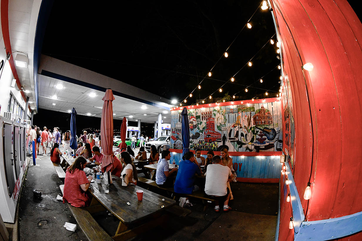 Customers sit at picnic tables outside 4 Corners Chevron at night, with string lights overhead and a large Oxford-themed mural on the wall.