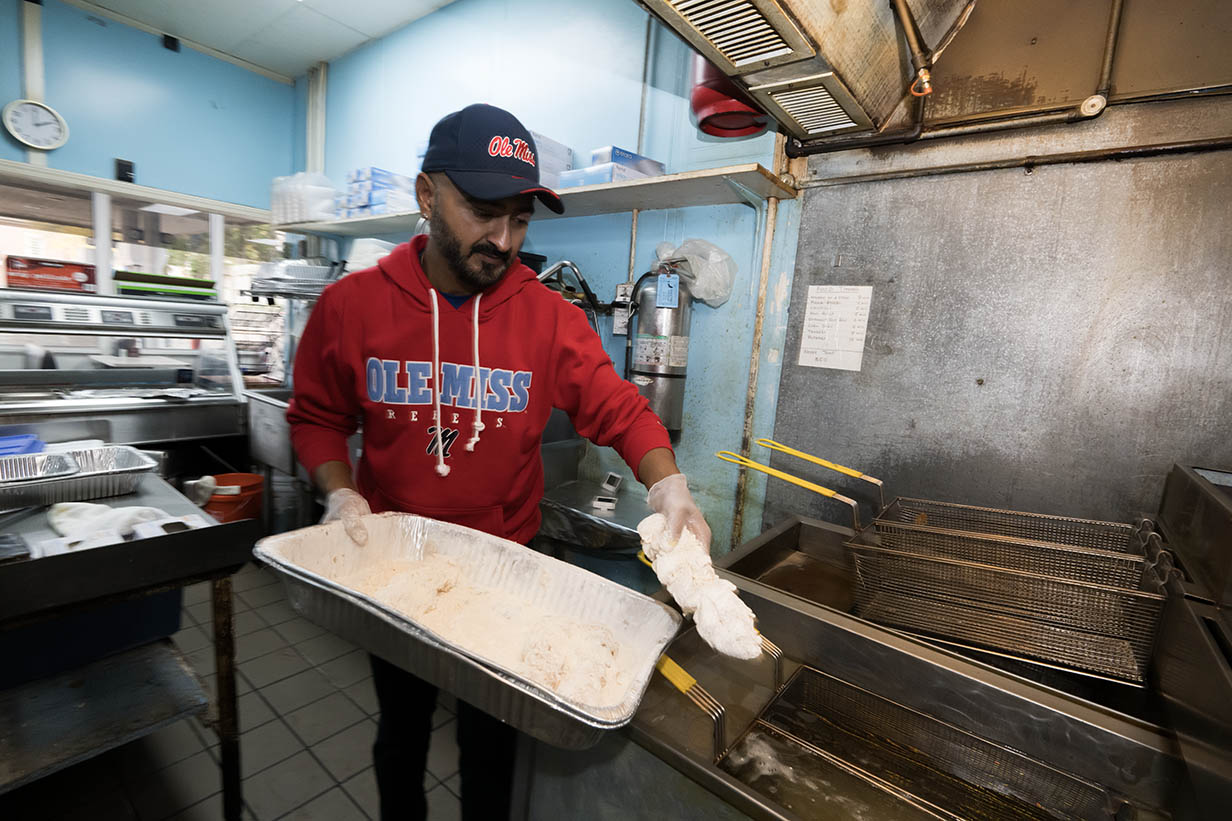 Owner Rafiq Rupani stands in the kitchen of 4 Corners Chevron, holding a tray of freshly battered chicken stick while working at a deep fryer.