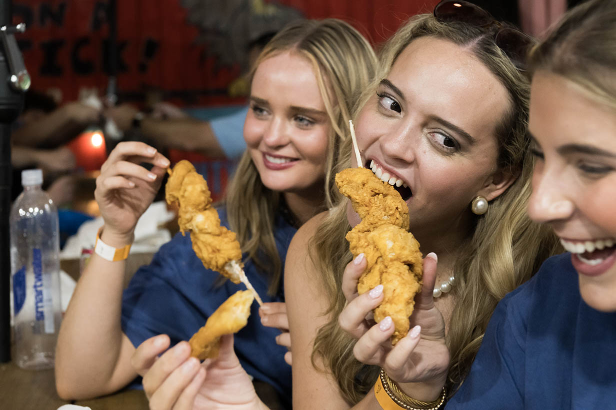 Three young women sit together smiling and holding pieces of chicken on a stick toward the camera.