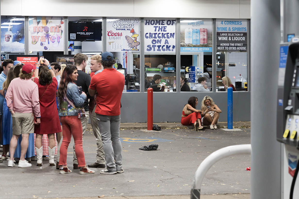 A group of people gathers outside the front windows of 4 Corners Chevron, some waiting in line and others sitting on the curb.