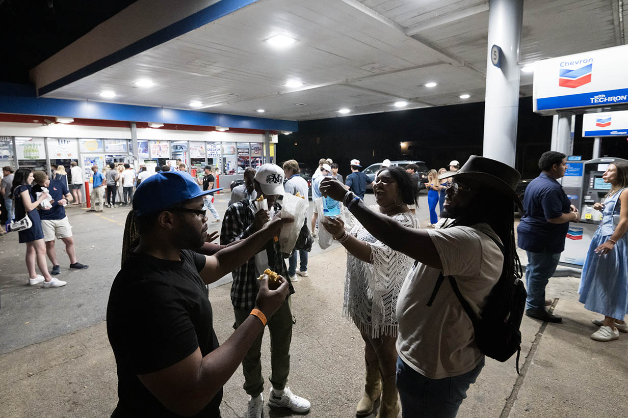 Several people stand outside 4 Corners Chevron at night, raising snacks and drinks together in a toast while other customers walk past.