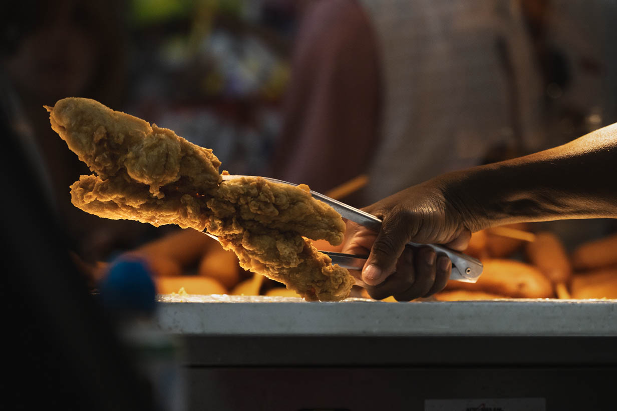 A hand holds two pieces of chicken on a stick over a warming counter at 4 Corners Chevron, lit by the heat lamps.