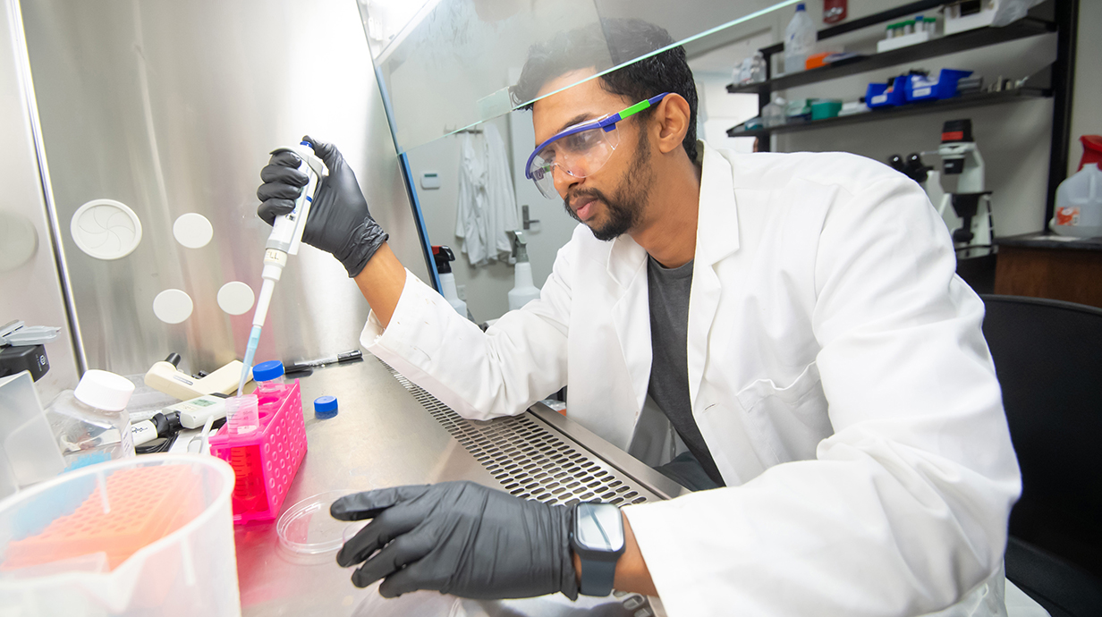 A young man wearing a white lab coat, goggles and gloves works with a pipette under a vented hood in a laboratory.