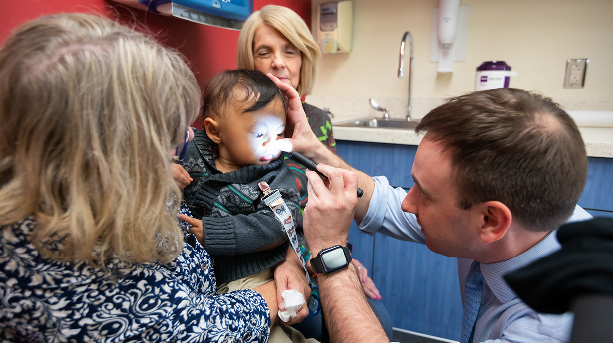 A doctor shines a small light at a child's mouth as two women watch.