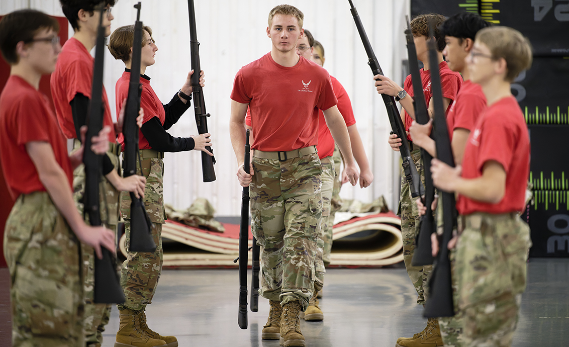 A young man wearing a red T-shirt and Army fatigues walks between two rows of young people dressed the same and holding drill rifles.