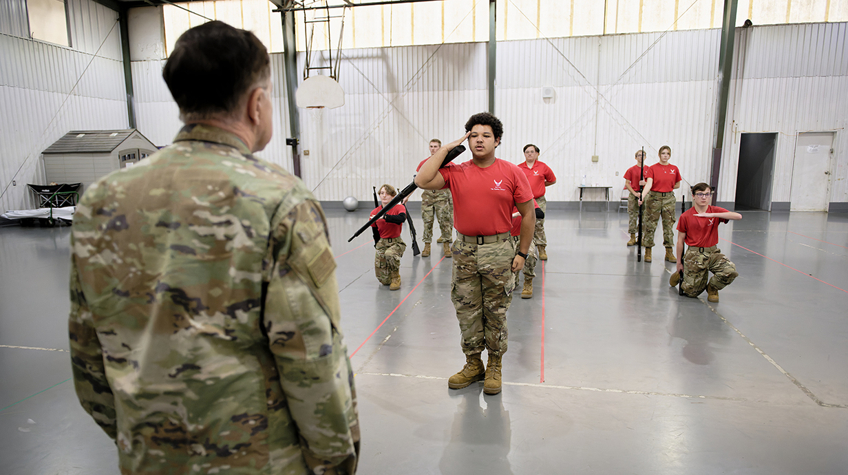 A young man wearing a red shirt and fatigues salutes a man wearing fatigues in a gym.