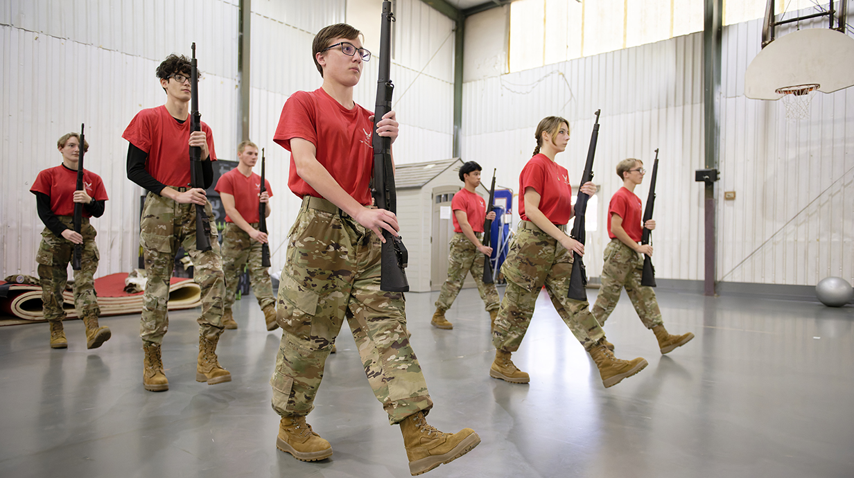 A group of young people wearing fatigues and red T-shirts practices marching in a gym.
