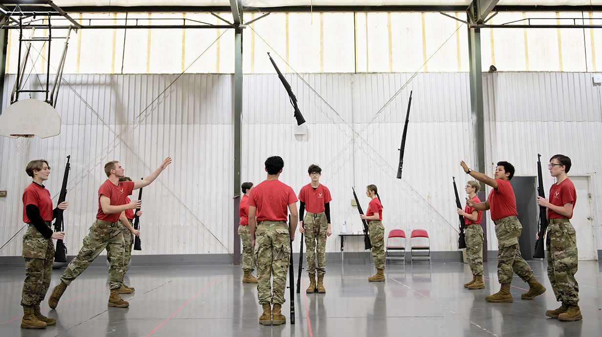 A group of young people wearing red T-shirts and fatigues practice tossing drill rifles in a gym.