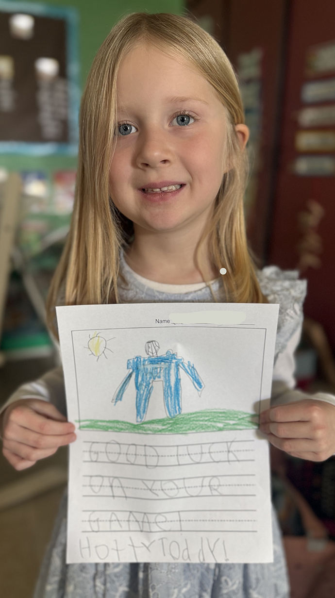 A young girl holds a drawing of a football player with a message written below it in pencil.
