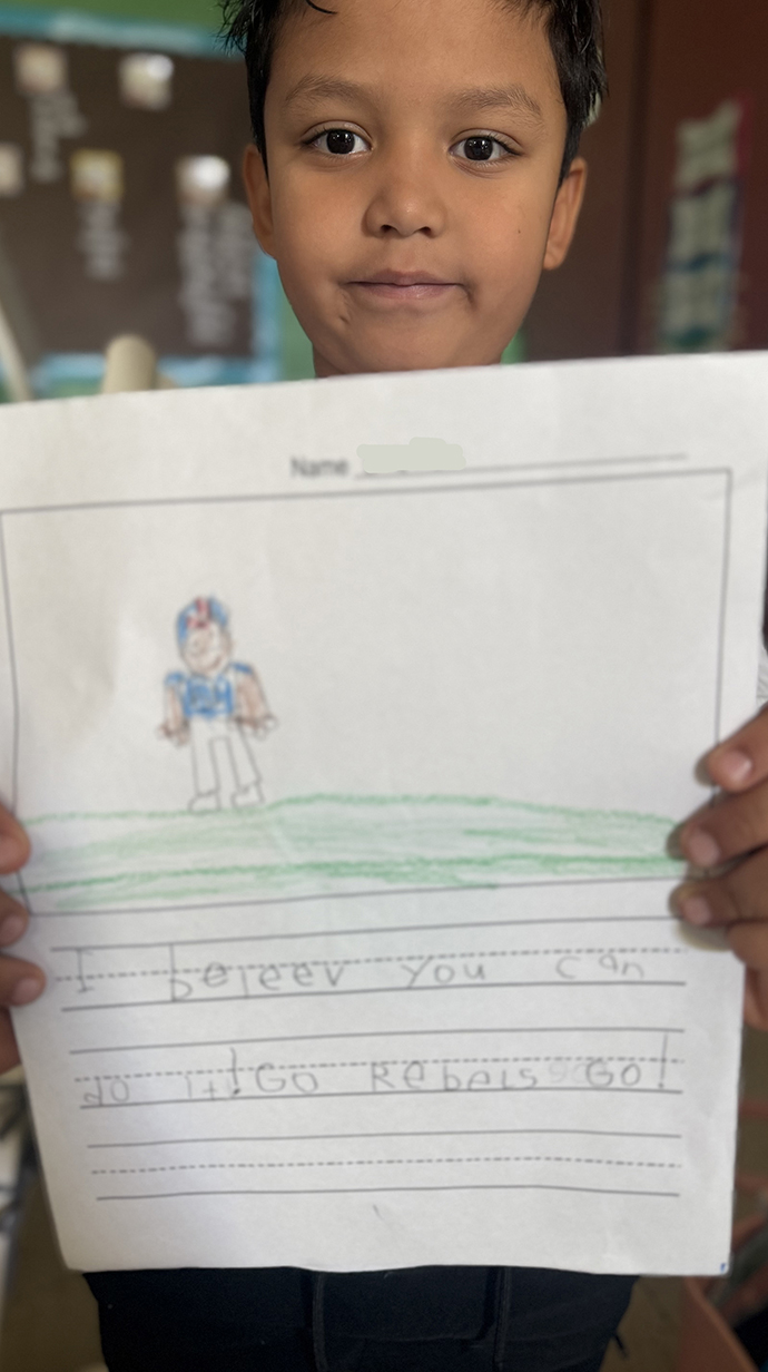 A young boy holds a drawing of a football player with a message written in pencil unerneath.