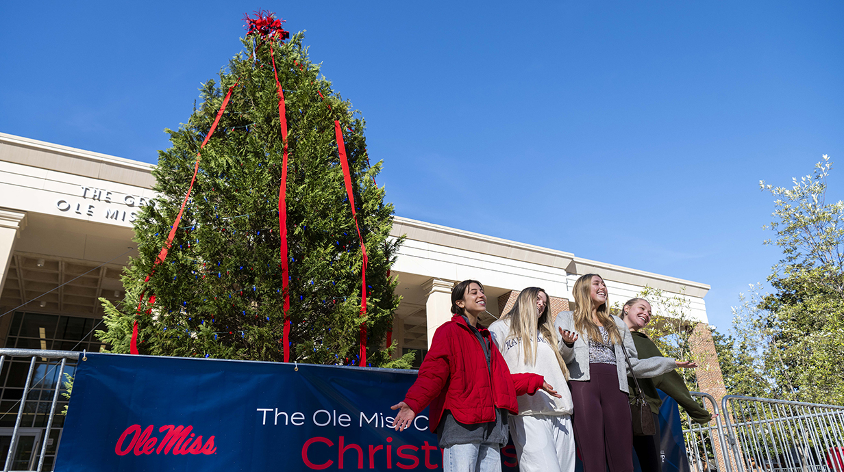 Four young women take a photo in front of a giant Christmas tree on an outdoor plaza.
