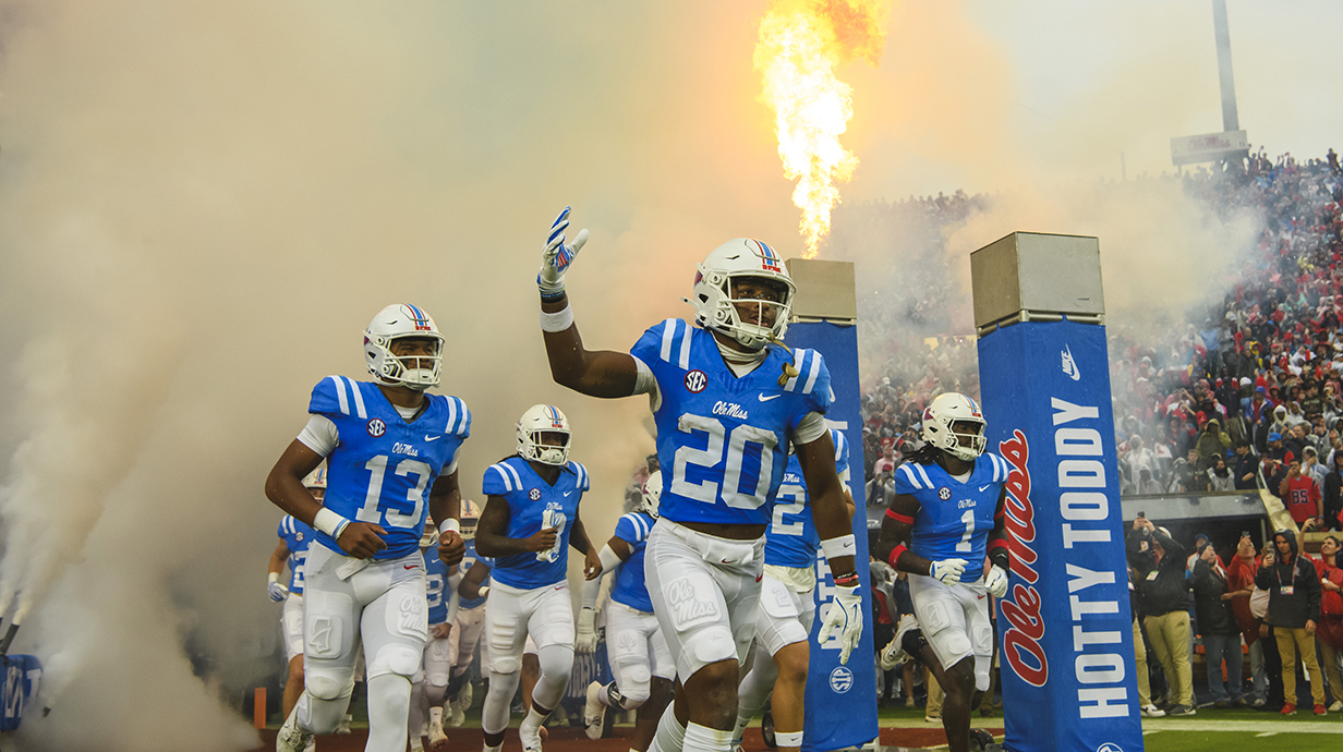 Ole Miss football players run onto the field at a stadium, surrounded by smoke and fire.