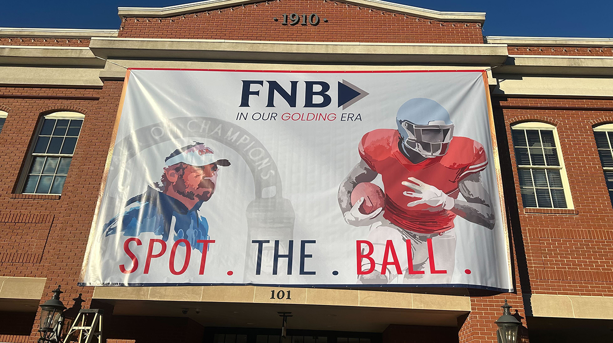 A banner reading 'Spot. The. Ball.' above photos of a man and a football player hangs on a red brick building.