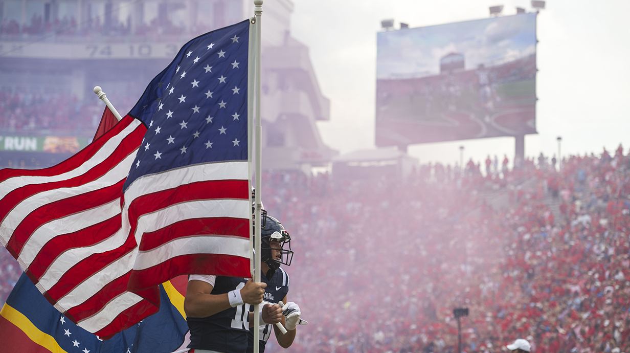 Football players carry an American flag and a Mississippi flag as they run into a crowded stadium.