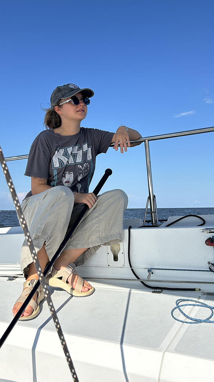 A young woman wearing sunglasses sits on the deck of a sailboat at sea.