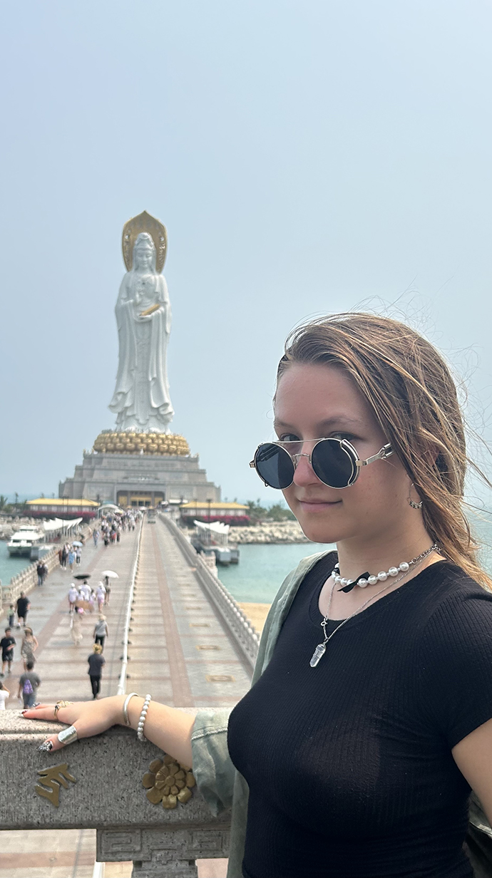A young woman looks over the top of her sunglasses while standing in front of a seaside statue.