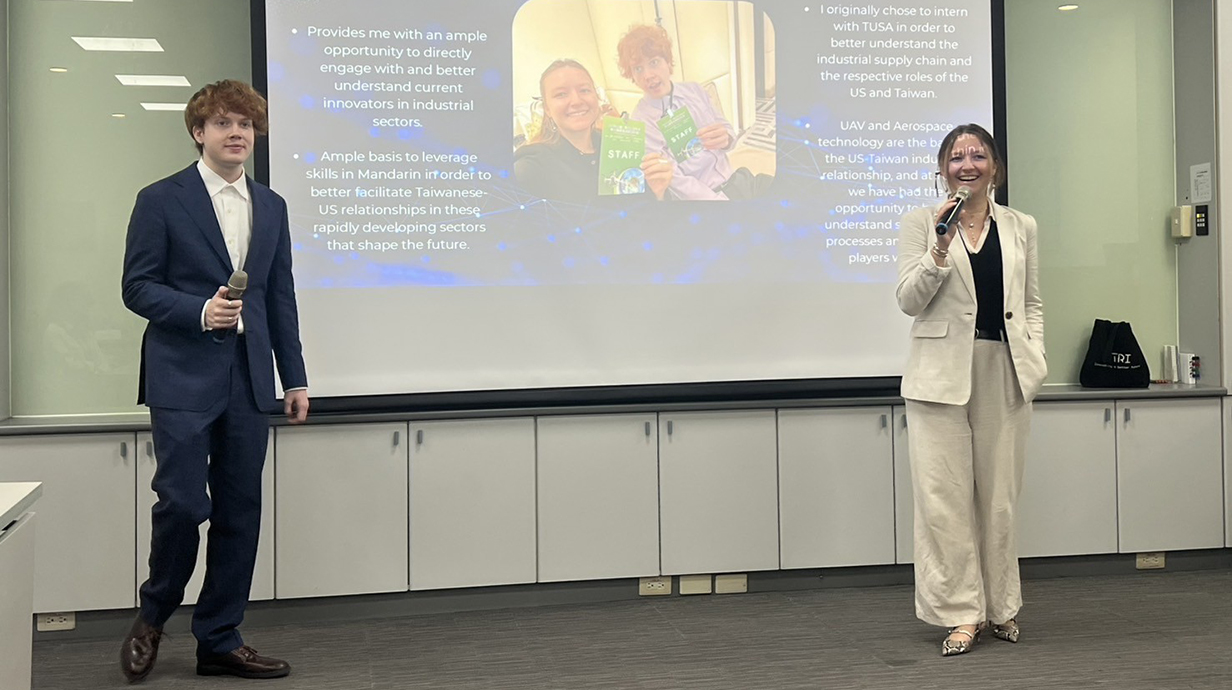 A young man and a young woman with microphones stand in front of a projection screen.