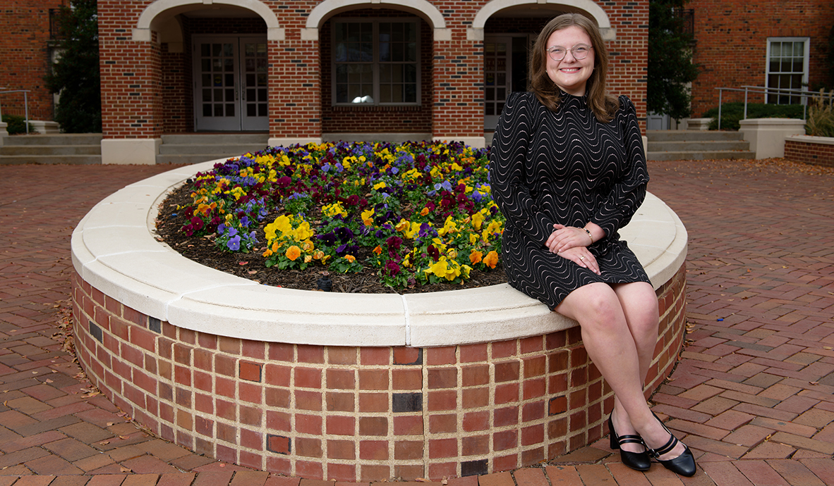 A young woman wearing a black dress sits on a brick planter full of flowers on a brick courtyard.