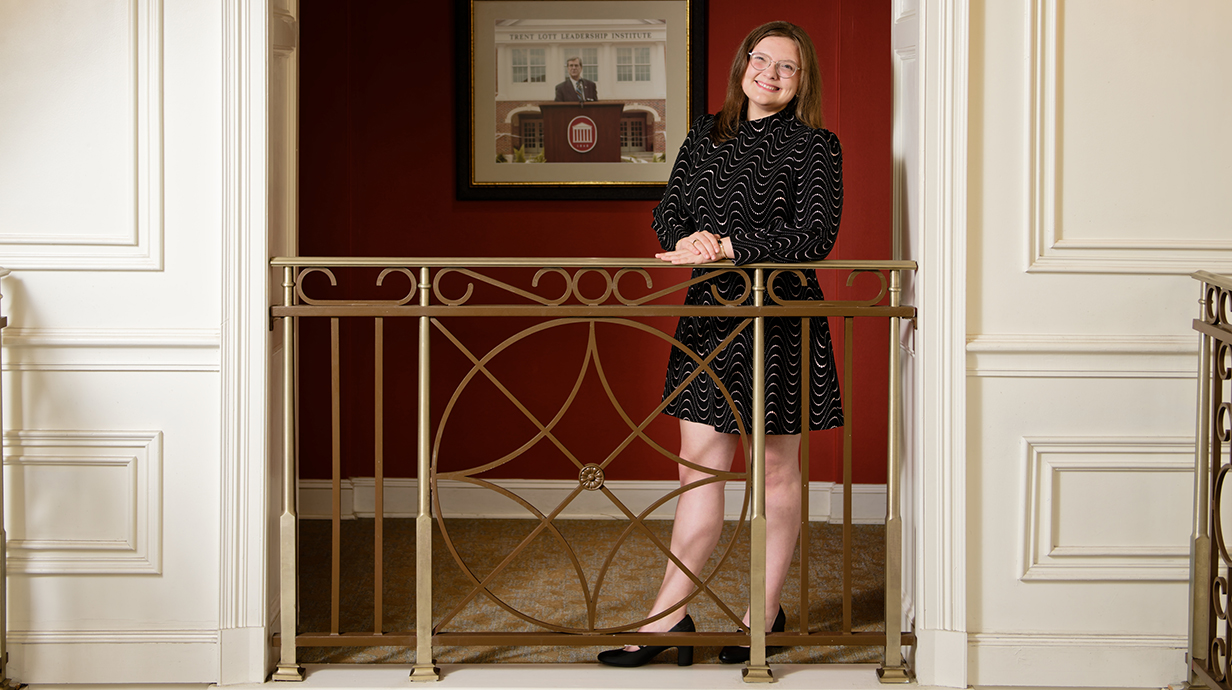 A young woman wearing a black dress stands at a railing on a second-floor landing.