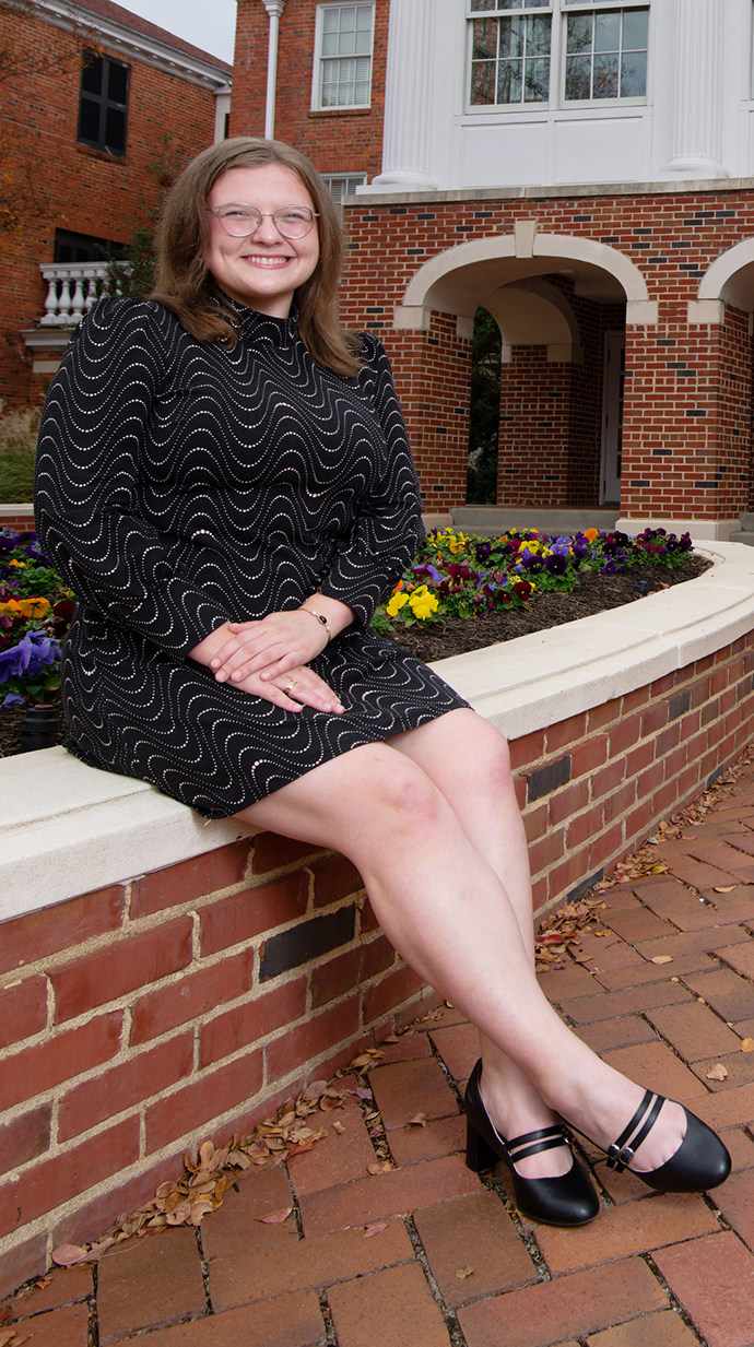 A young woman wearingn a black dress sits on a brick planter full of colorful flowers.