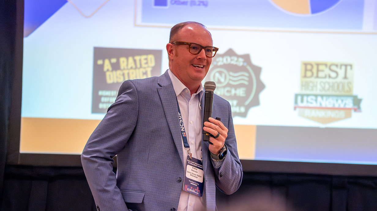 A man wearing a suit with no tie speaks into a microphone in front of a projection screen.