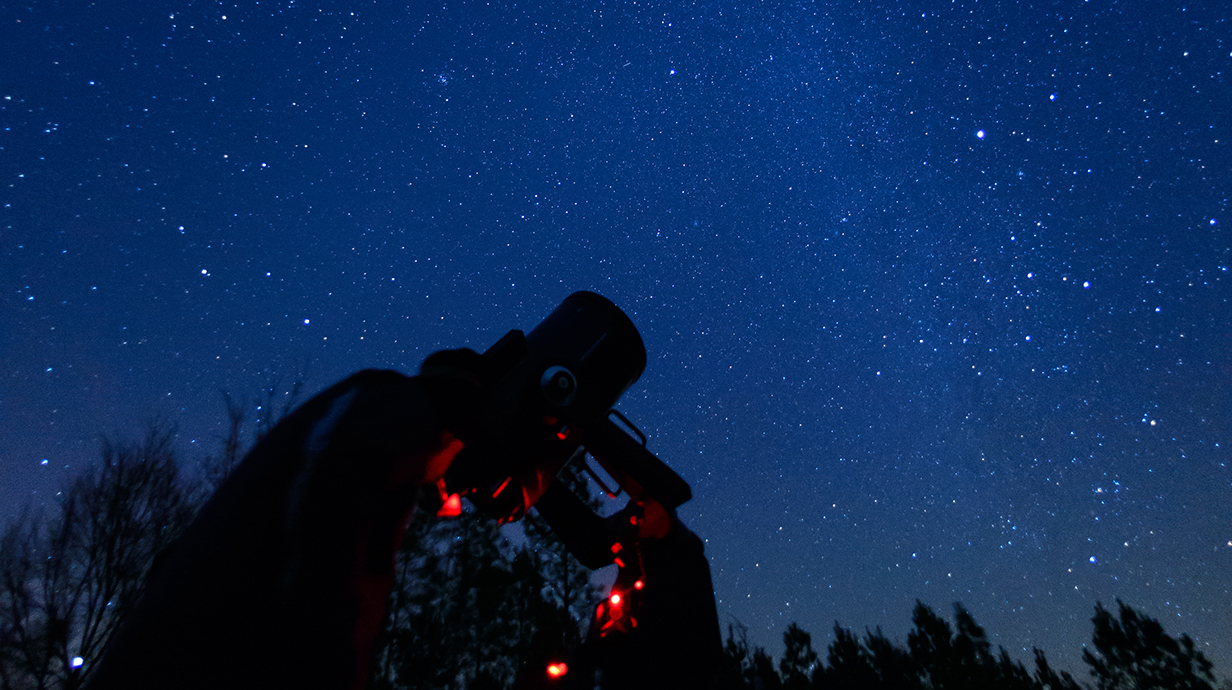 A person looking through the eyepiece of a telescope is silhouetted against a starry night sky.