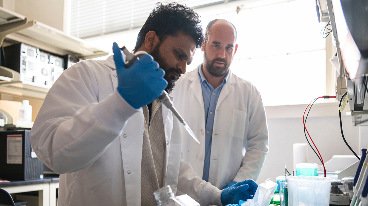 A young man uses a pipette to transfer liquid in a laboratory while another man watches.