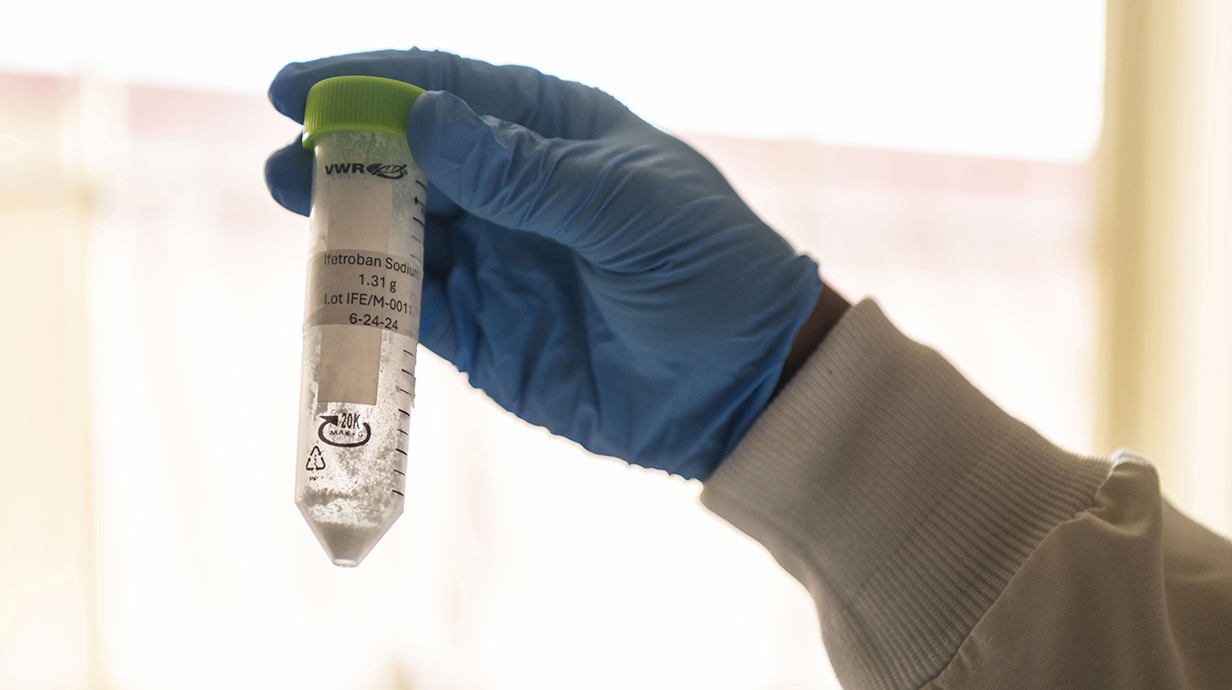 A hand wearing a blue latex glove holds a vial of liquid in a laboratory.