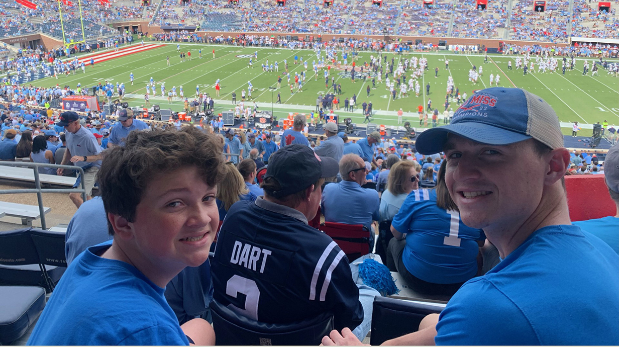 Two young men sitting in a crowded football stadium look over their shoulders at the camera.