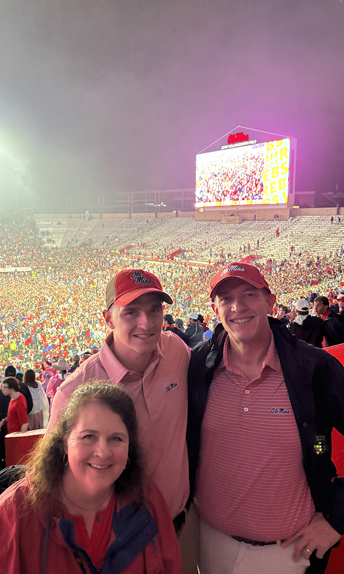A woman and two men pose together in a crowded football stadium after a game.