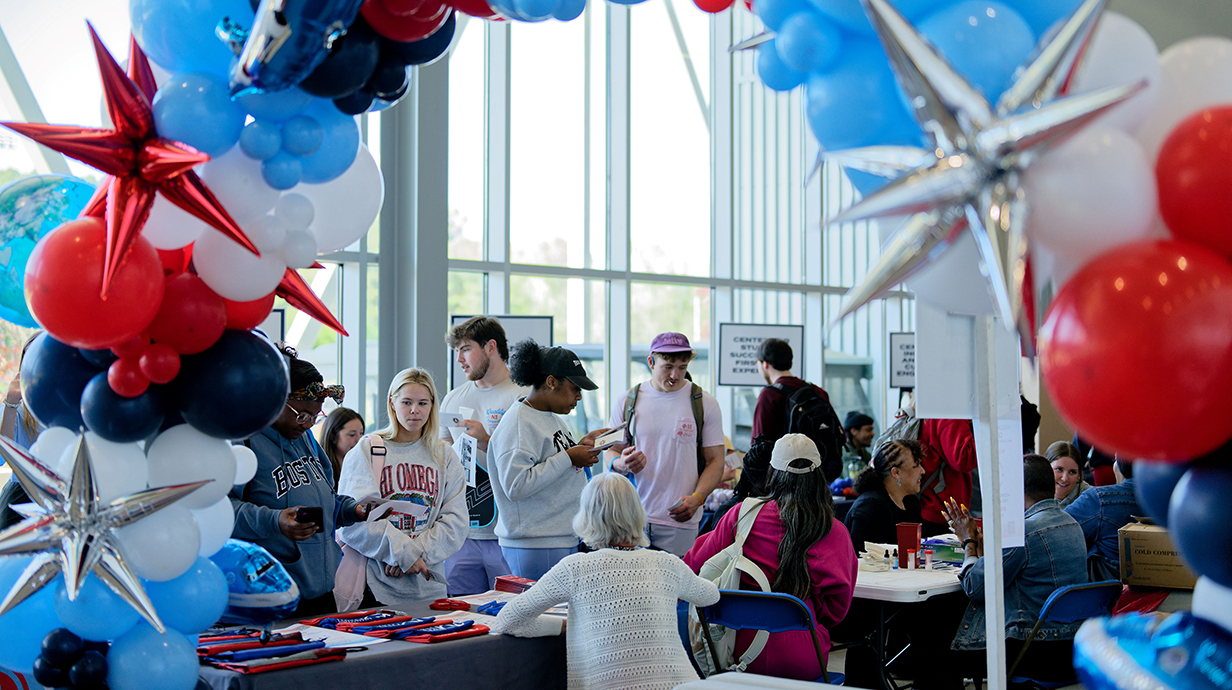 A group of young people look over items  displayed on a table topped by an arch of red, while and blue balloons.