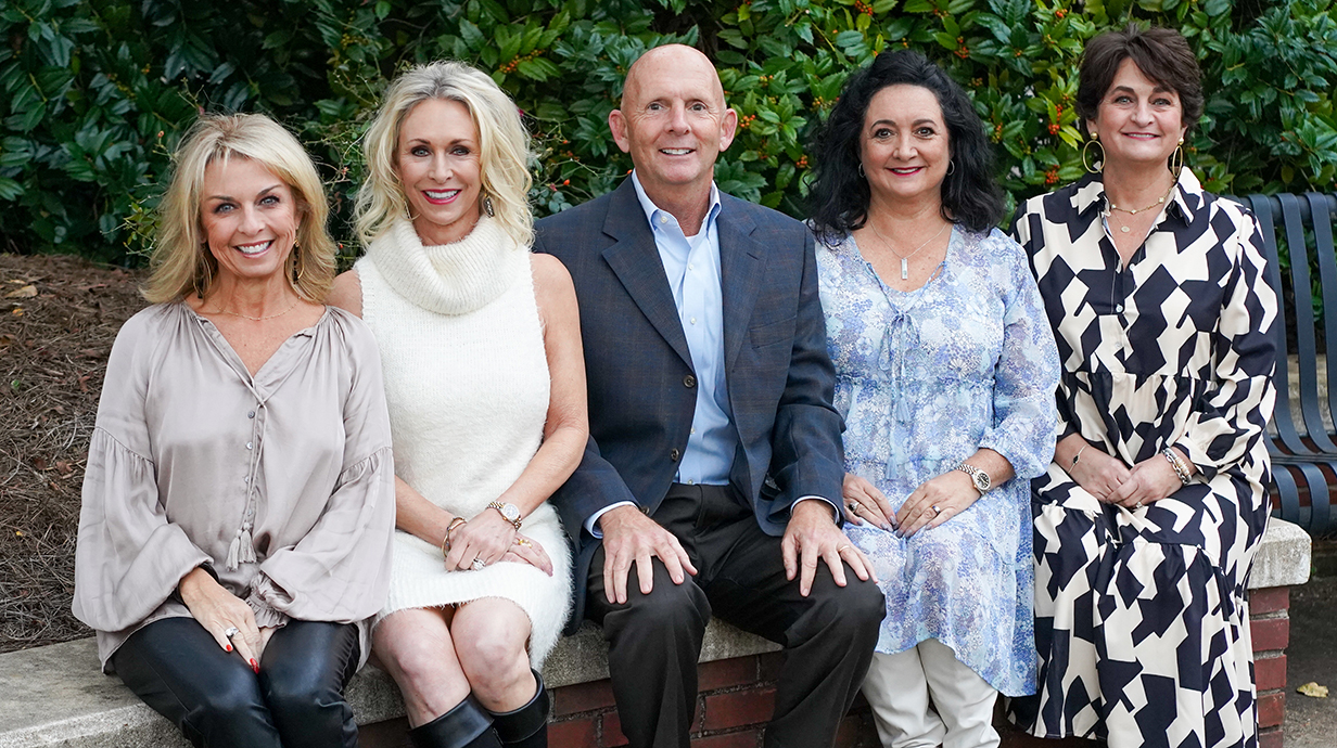Four women and a man sit on a low brick wall surrounding a landscaped area.
